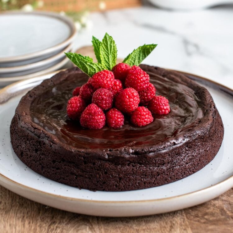 a flourless chocolate cake on a serving platter. The cake is topped with raspberries and mint leaves.