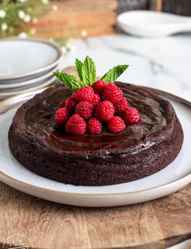 a flourless chocolate cake on a serving platter. The cake is topped with raspberries and mint leaves.