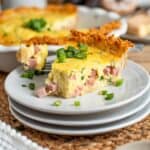 a slice of ham and cheese quiche with a crispy potato hash brown crust, garnished with sliced green onions. The quiche is on a small white plate on a table.