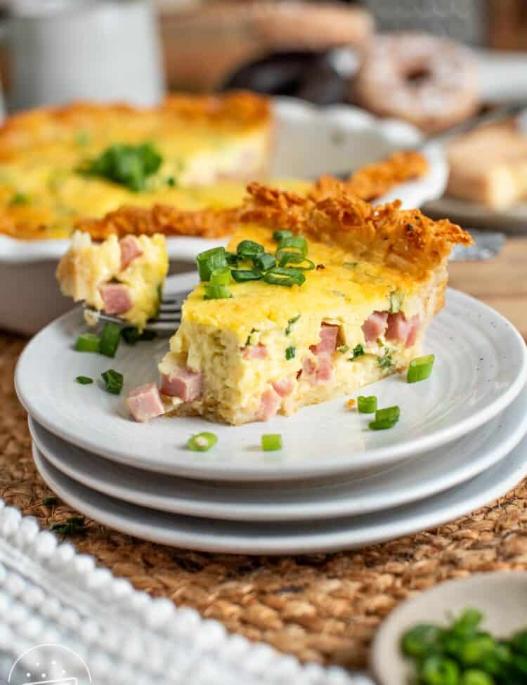 a slice of ham and cheese quiche with a crispy potato hash brown crust, garnished with sliced green onions. The quiche is on a small white plate on a table.