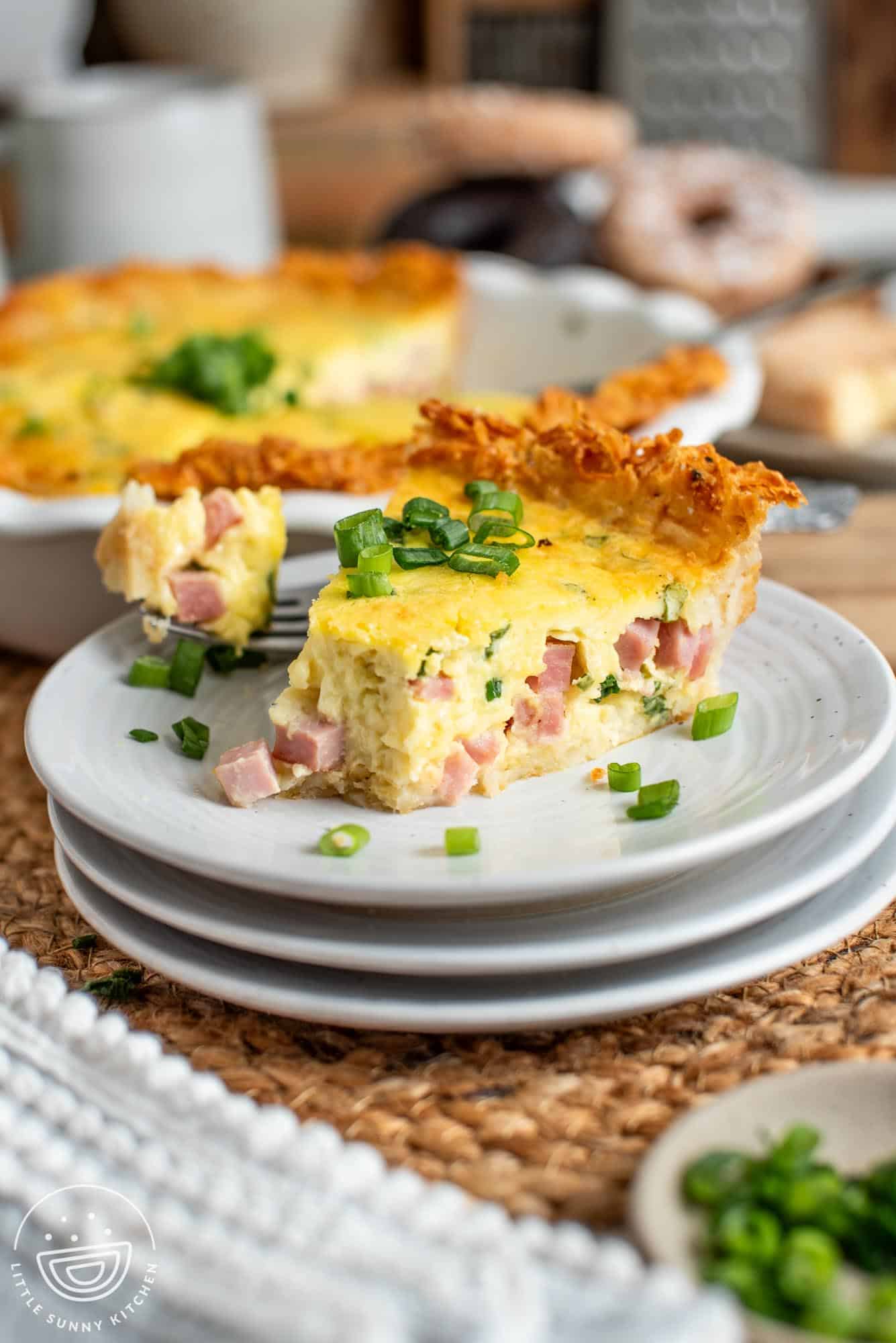 a slice of ham and cheese quiche with a crispy potato hash brown crust, garnished with sliced green onions. The quiche is on a small white plate on a table.