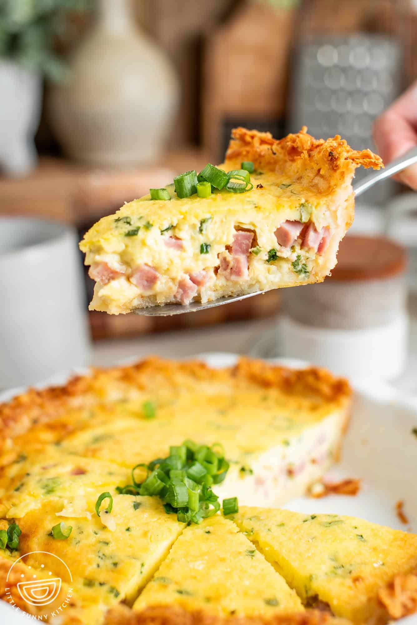 a spatula lifting up a slice of hash brown crust quiche from the whole pie. The texture of the quiche is light and creamy with diced ham and green onions visible.