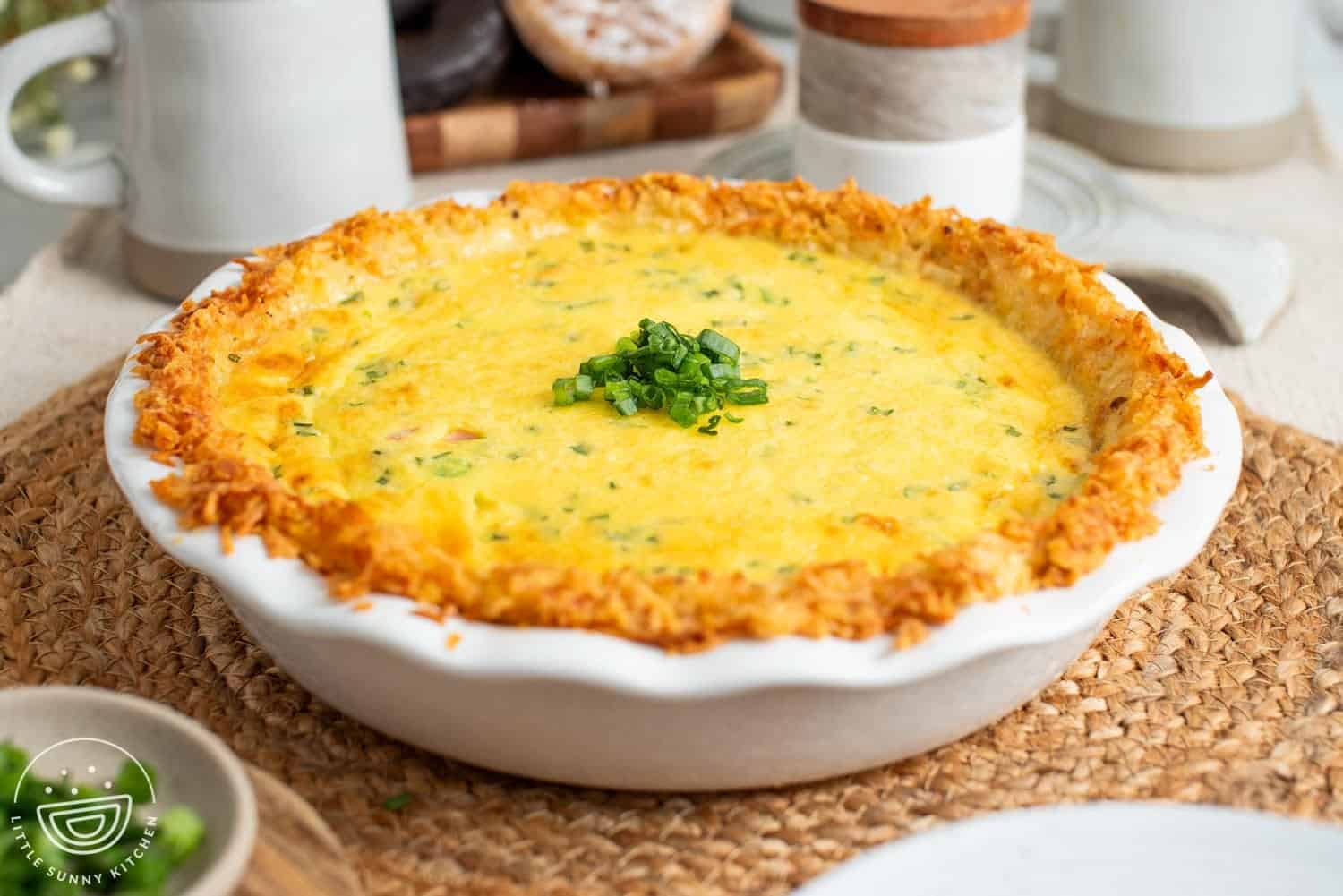 a quiche in a white ceramic pie plate with a crispy hash brown crust. The pie is sitting on a jute place mat.