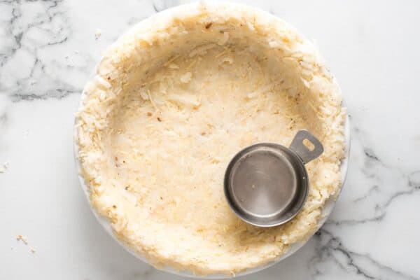 Hash brown crust for quiche pressed into a pie pan with a small metal measuring cup.