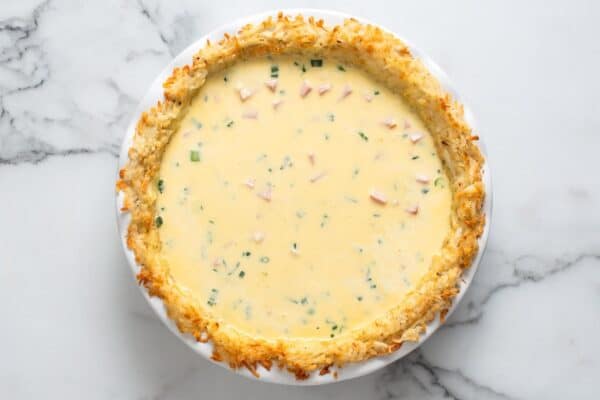ham and cheese quiche mixture poured into a baked hash brown crust that is sitting on a marble counter.