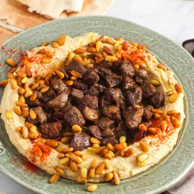 Hummus served on a green plate, topped with cooked diced meat and toasted pine nuts. With pita bread in the background.