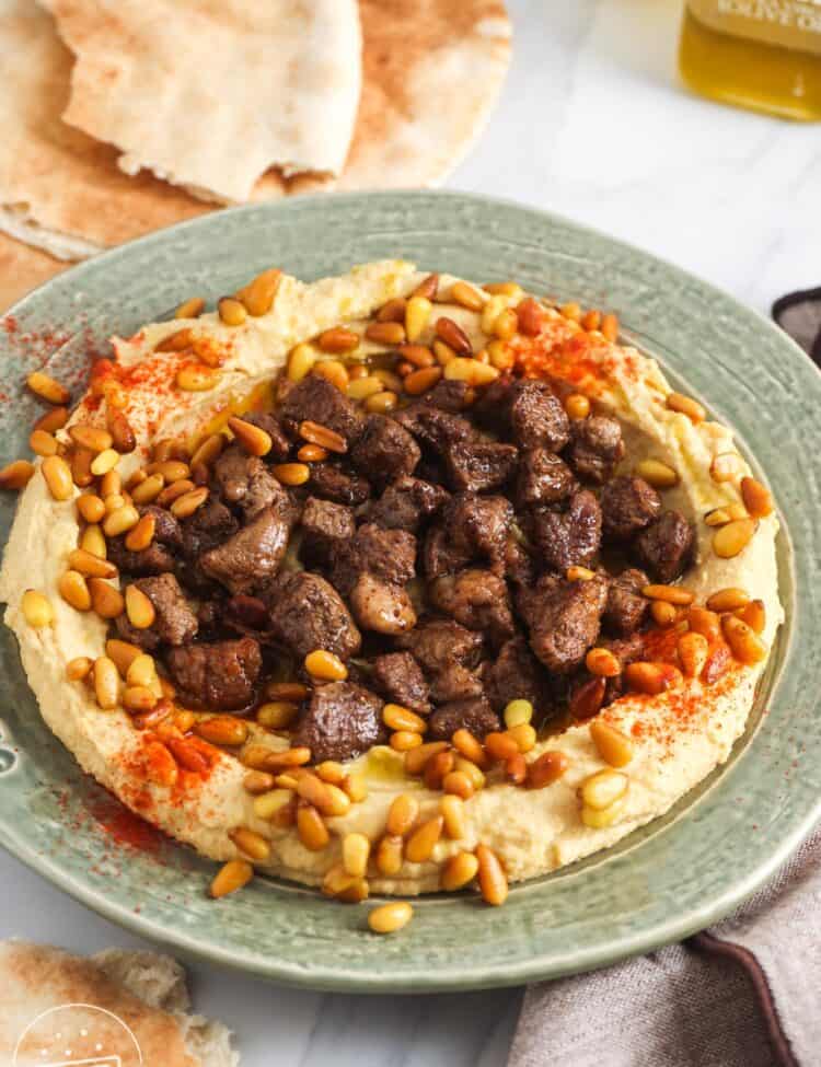 Hummus served on a green plate, topped with cooked diced meat and toasted pine nuts. With pita bread in the background.