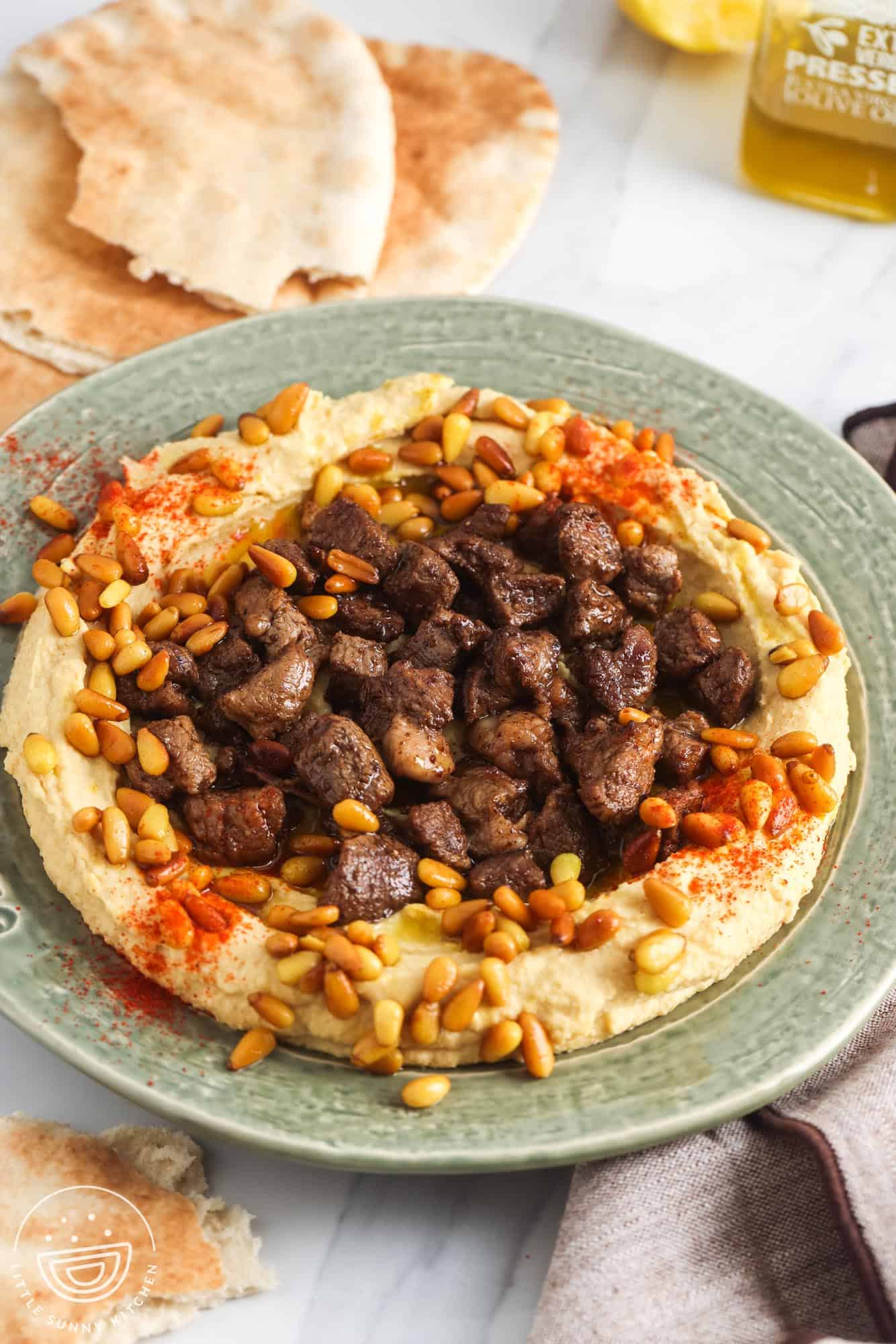 Hummus served on a green plate, topped with cooked diced meat and toasted pine nuts. With pita bread in the background.