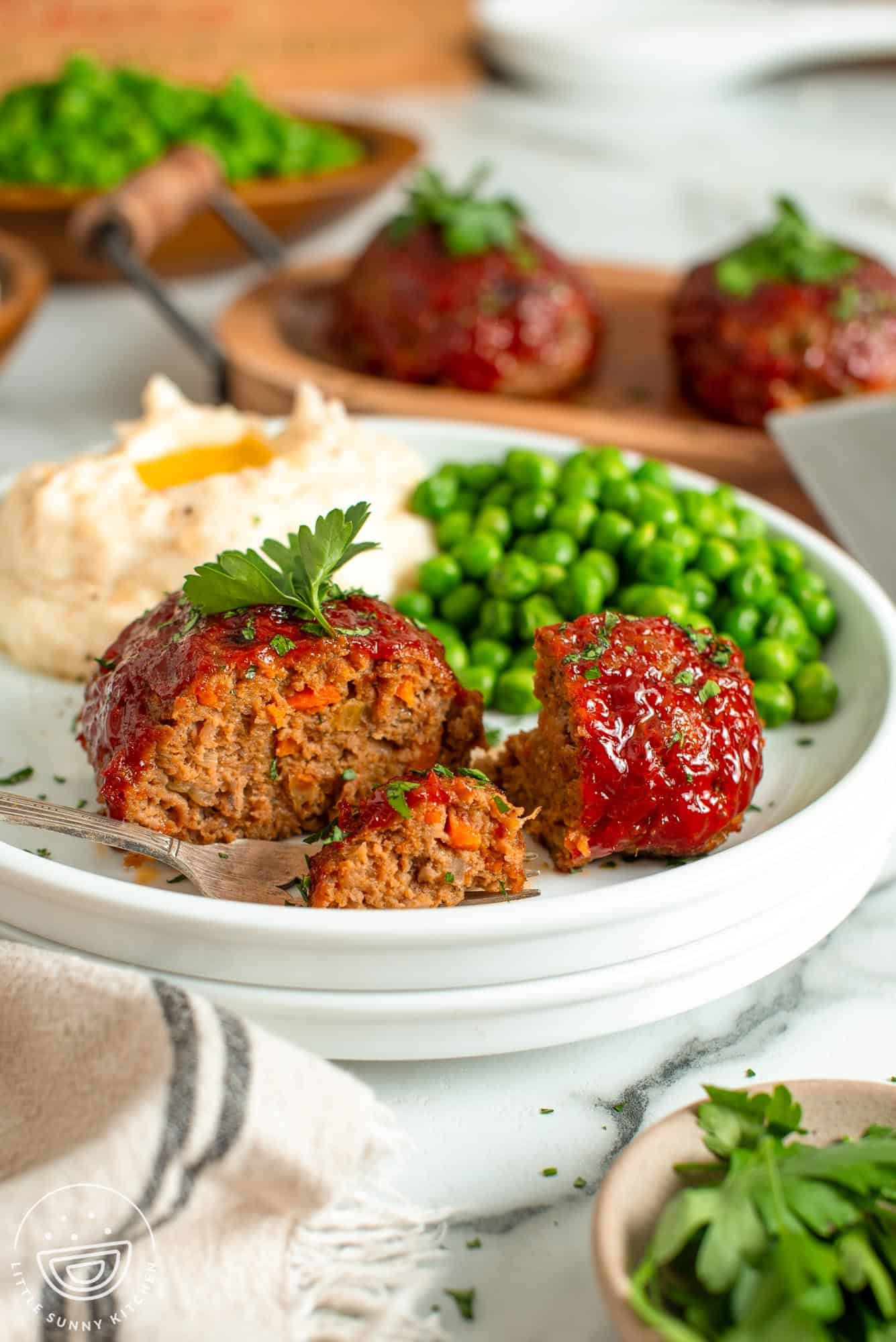 a white dinner plate with one mini meatloaf patty, green peas, and mashed potatoes.
