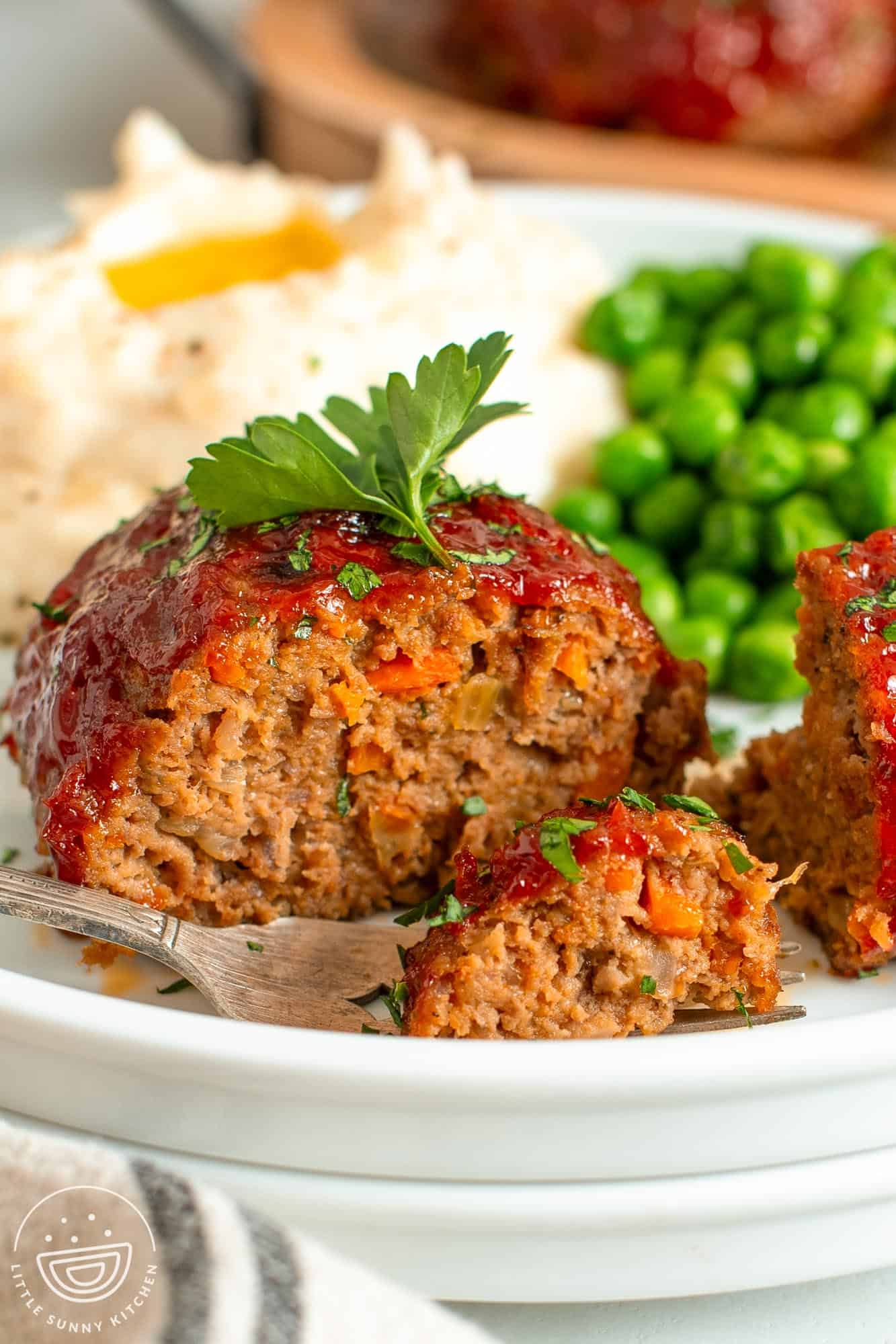 closeup of a mini meatloaf on a dinner plate with peas and potatoes. The meatloaf has been cut in half to show the moist texture.
