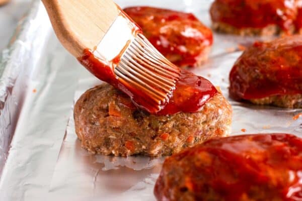 a pastry brush adding red ketchup glaze to uncooked mini meatloaves.