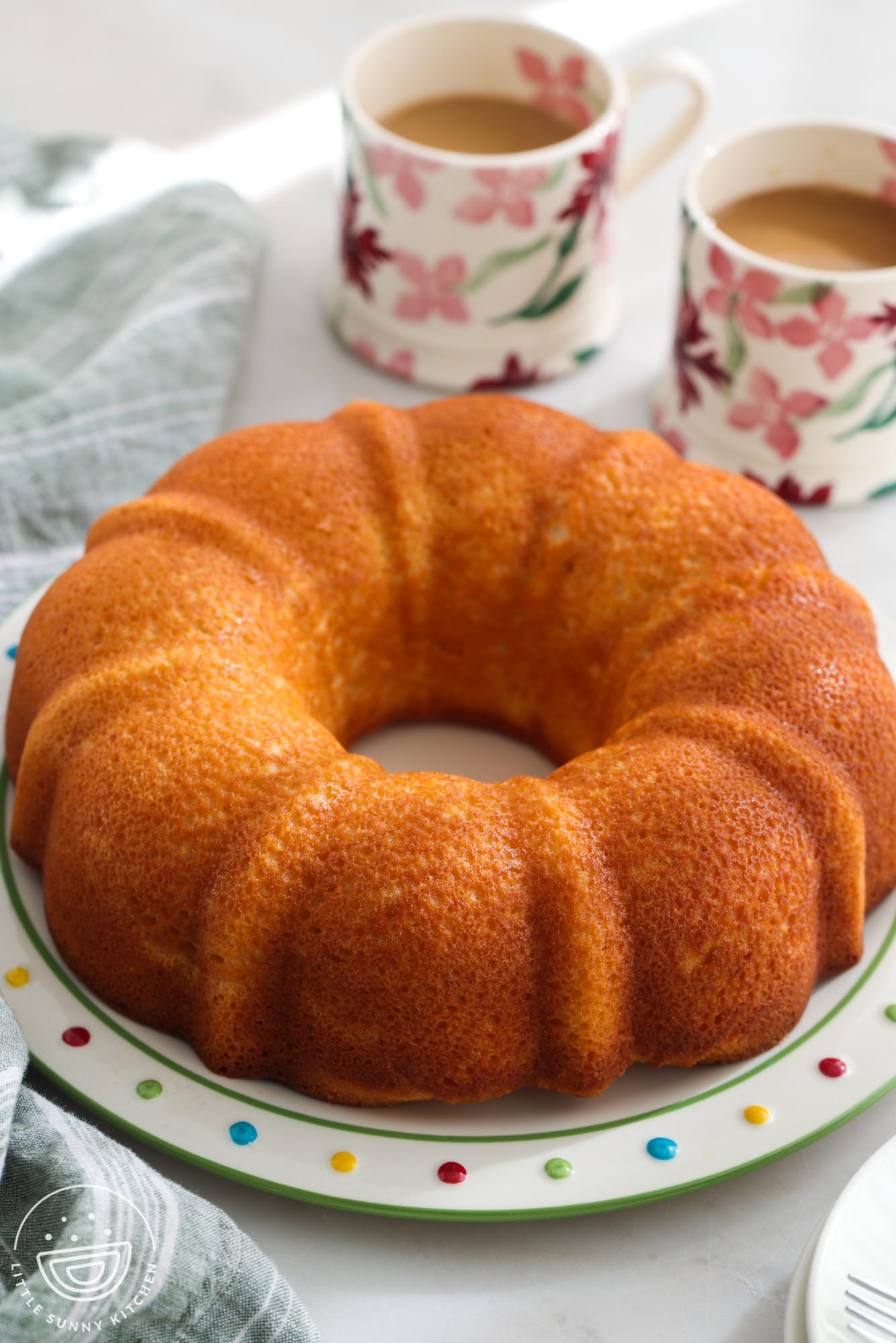 a whole yogurt pound cake, baked in a bundt pan, on a colorful polka dot plate. Next to the cake are two floral mugs of coffee.