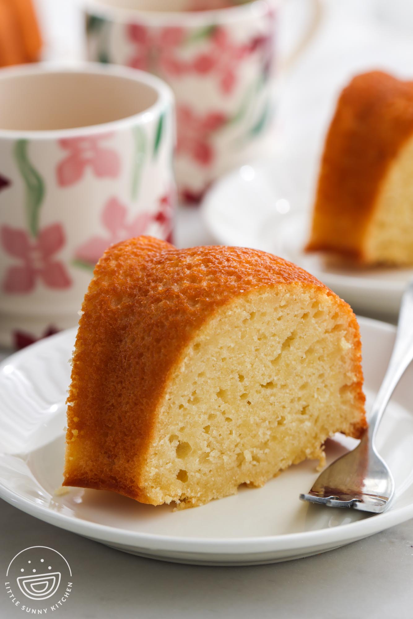 a slice of yogurt bundt cake on a small plate with a fork. In the background are floral coffee mugs.