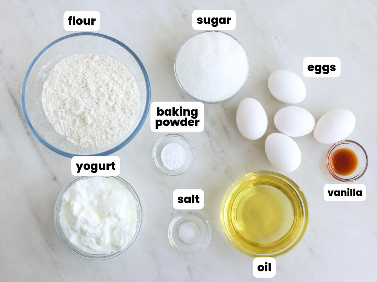 Ingredients to make yogurt pound cake, including flour, sugar, yogurt, baking powder, salt, oil, eggs, and vanilla, on a marble counter.
