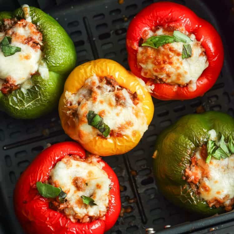 5 stuffed peppers (2 red, two green, and one yellow) in the basket of a square air fryer.