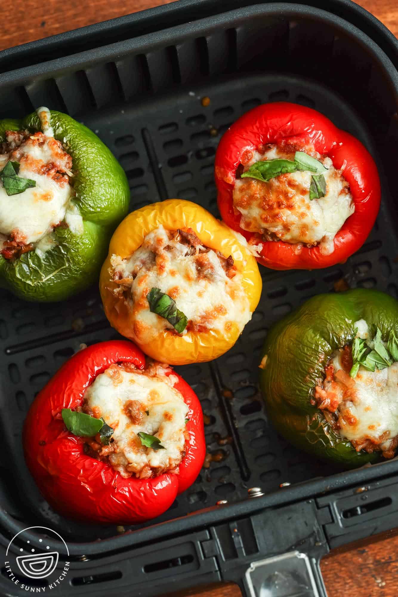 5 stuffed peppers (2 red, two green, and one yellow) in the basket of a square air fryer.