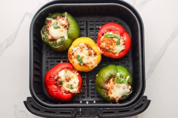 5 stuffed peppers in an air fryer, topped with melted, browned cheese.