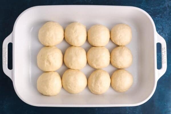 12 unbaked dinner rolls in a white ceramic baking dish. The rolls are touching each other.