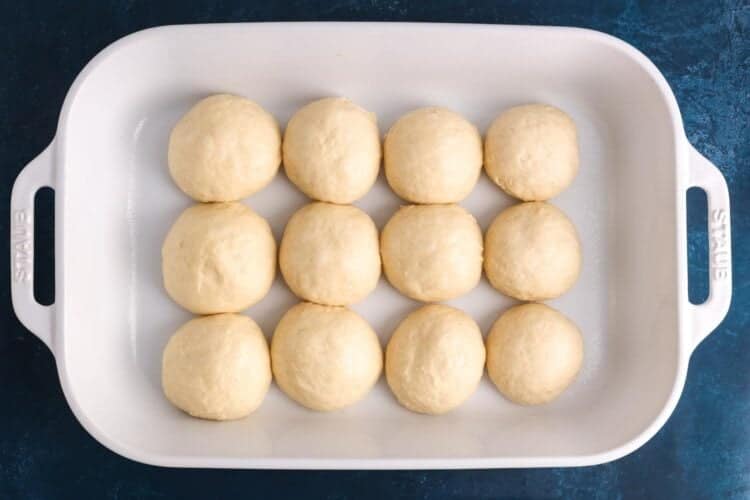 12 unbaked dinner rolls in a white ceramic baking dish. The rolls are touching each other.