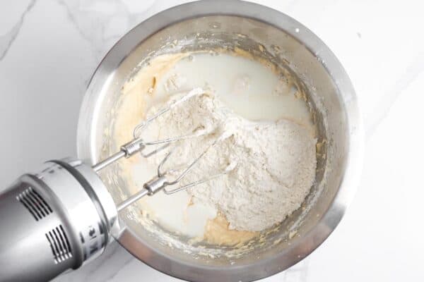 Milk and flour added to muffin batter in a metal bowl with an electric hand mixer.
