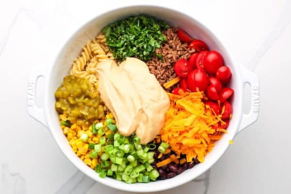 a large white bowl filled with pasta, veggies, cheese, ground beef, and topped with a creamy dressing.