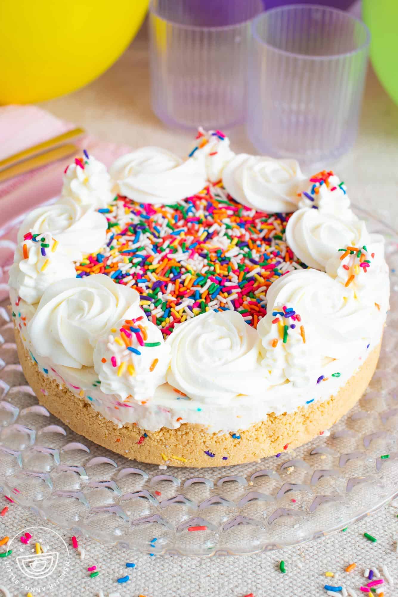 large rosettes of whipped cream top a funfetti cheesecake with a golden cookie crust. The cheesecake is on a glass platter with party balloons in the background.