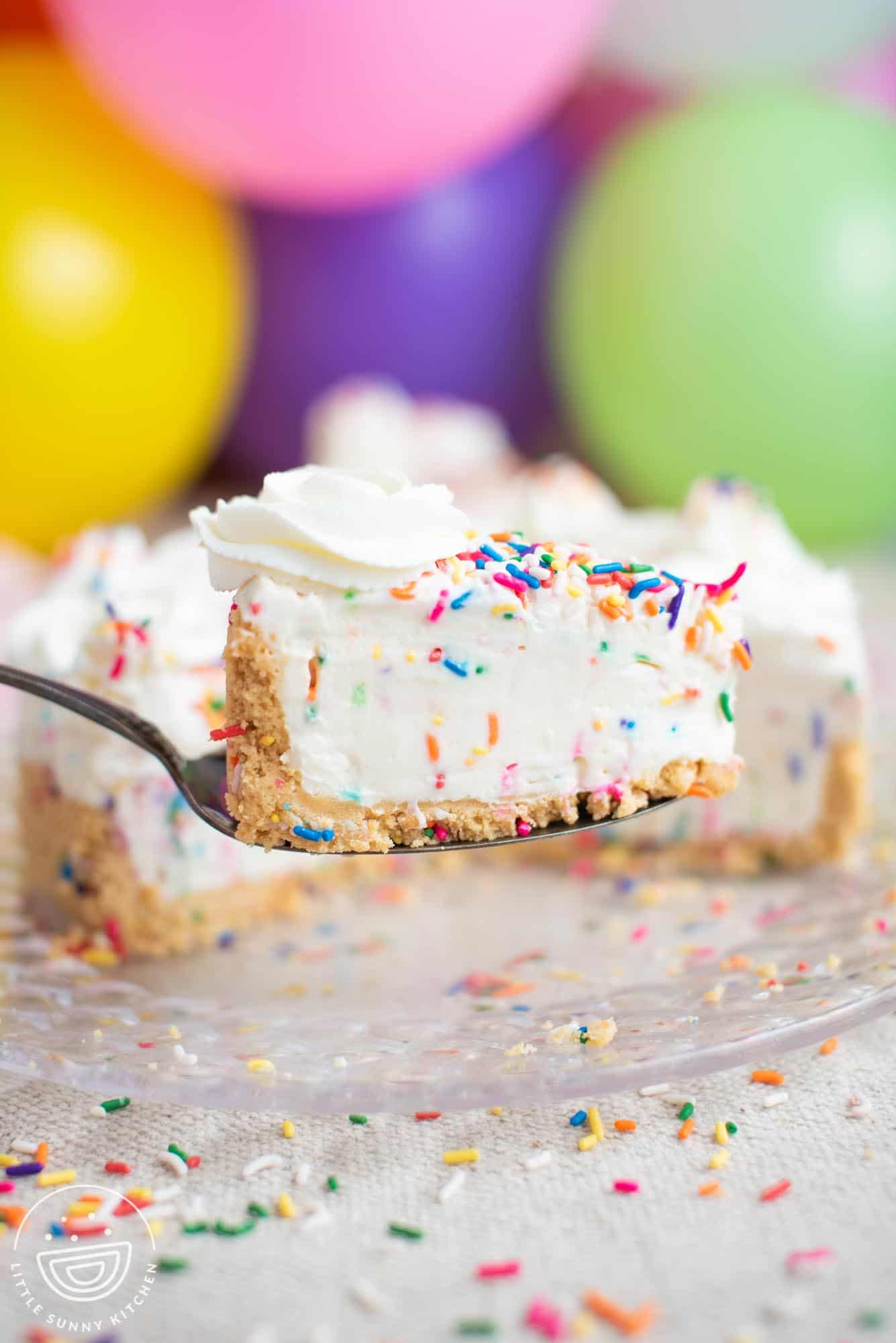 a spatula holding up a slice of no bake funfetti cheesecake filled with sprinkles. party balloons are in the background.