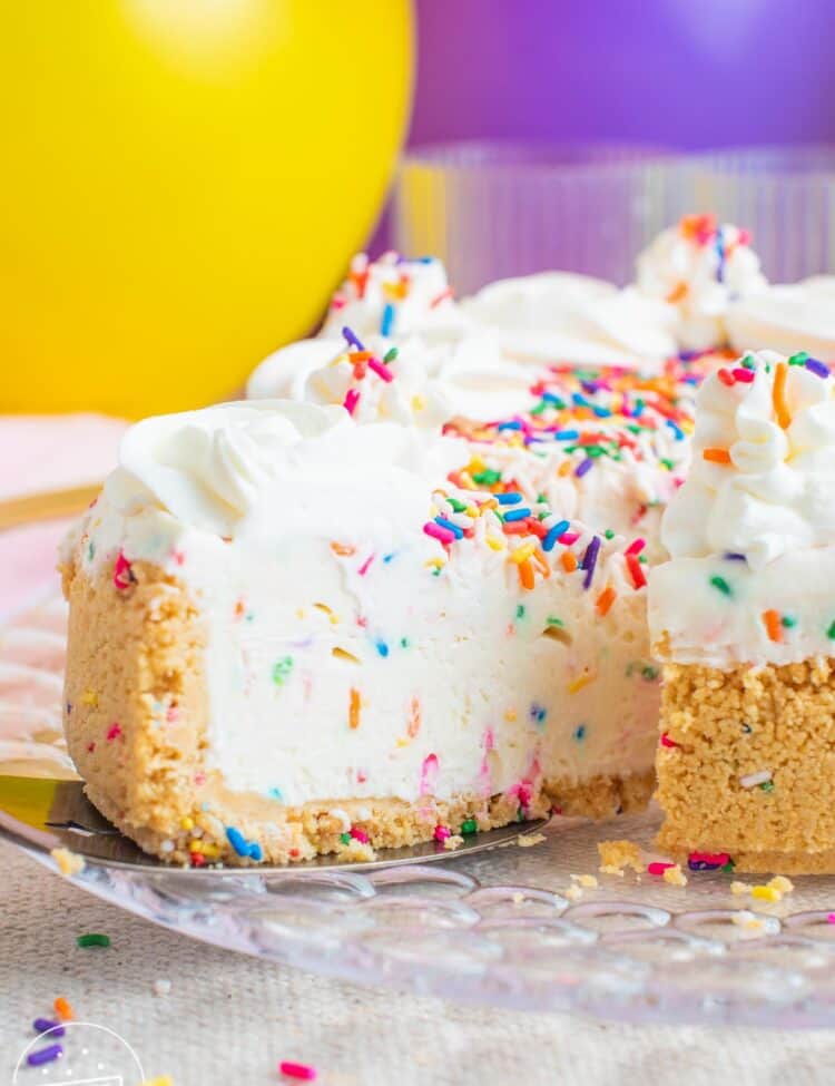 a slice of funfetti cheesecake being removed from the whole pie. In the background is a yellow and a purple balloon.