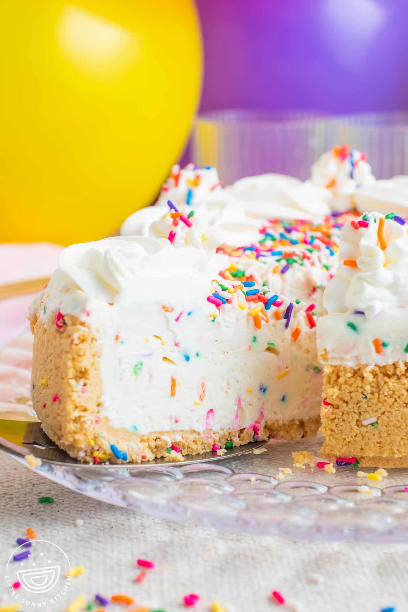 a slice of funfetti cheesecake being removed from the whole pie. In the background is a yellow and a purple balloon.