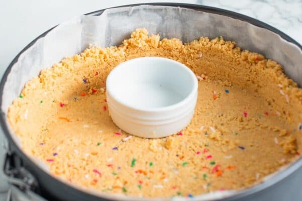 Golden Oreo cookie crust pressed into a springform pan that is lined with parchment paper. a white ramekin is used to make the crust even and level.