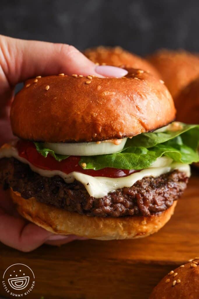 a hand holding a cheeseburger with toppings that is on a homemade toasted hamburger bun.