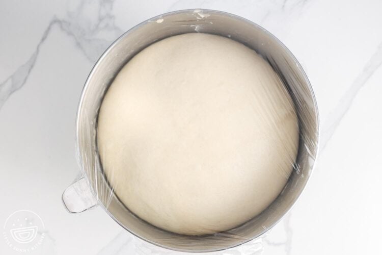 Risen dough in a metal bowl that is covered with plastic wrap.