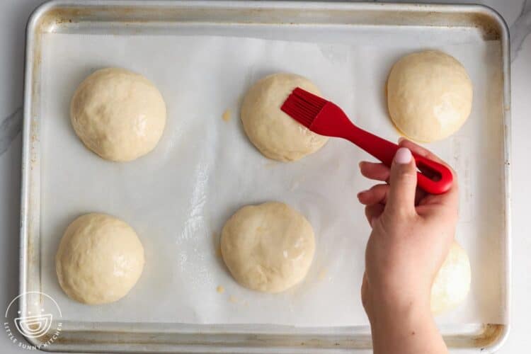a hand using a red brush to add egg wash to the top of dough for buns.