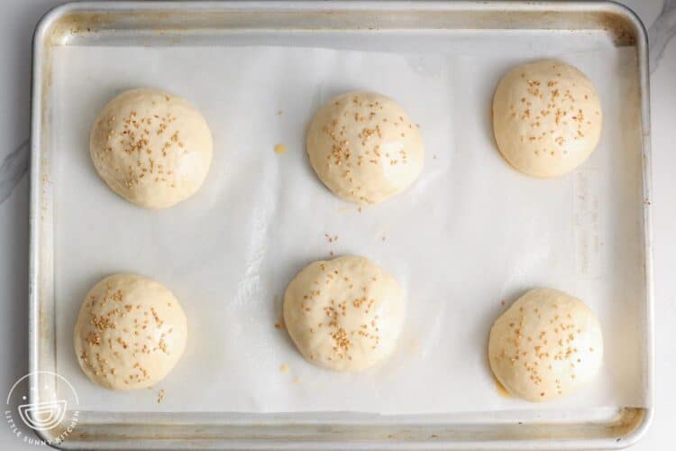 unbaked hamburger buns on parchment. The buns have been sprinkled with sesame seeds.