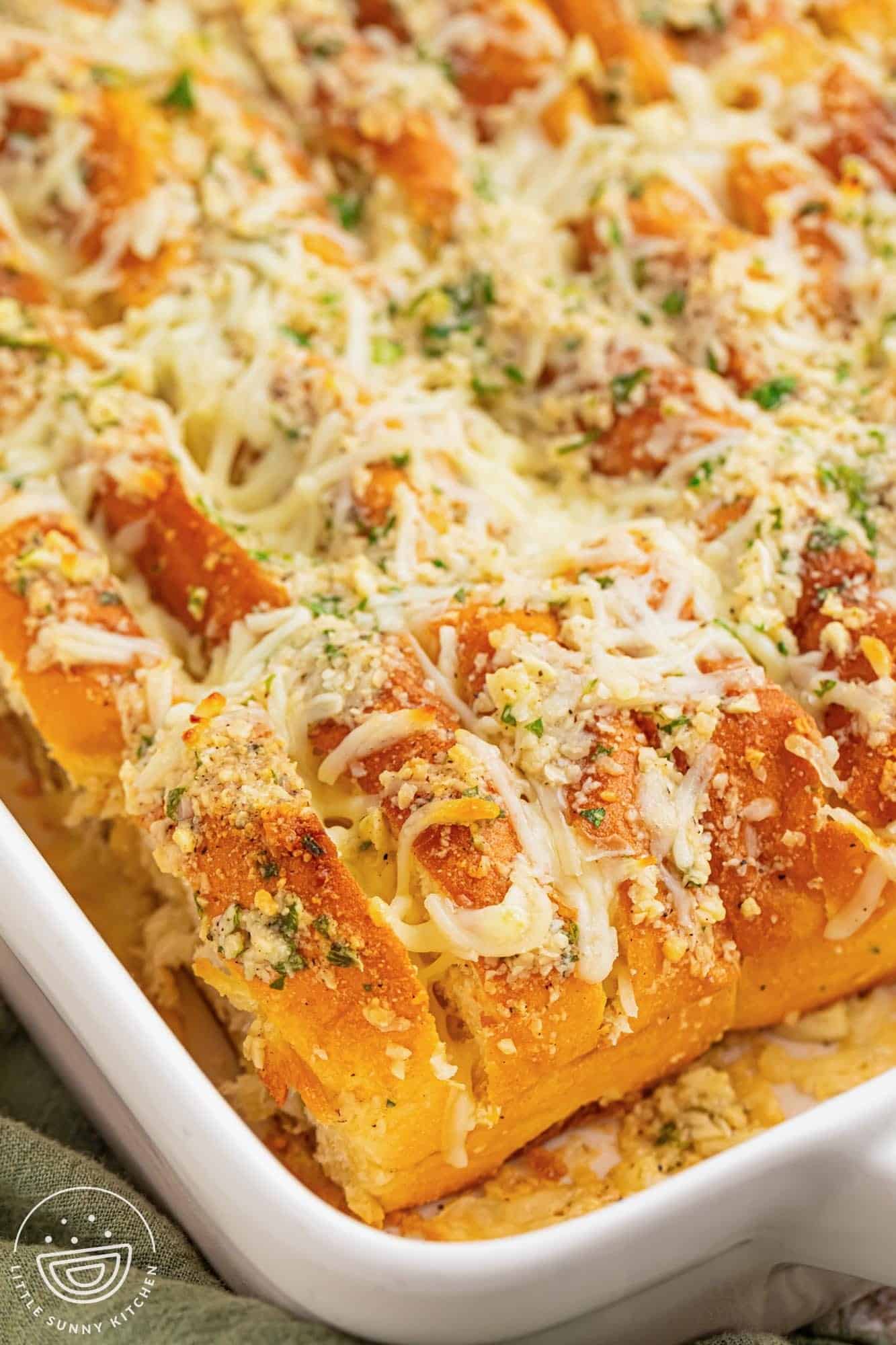 sliced hawaiian roll garlic bread in a white ceramic baking dish. The buns are covered in cheesy garlic butter and chopped herbs.