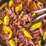 shredded Mississippi pot roast in a skillet with pepperoncini and gravy. Tongs are serving.