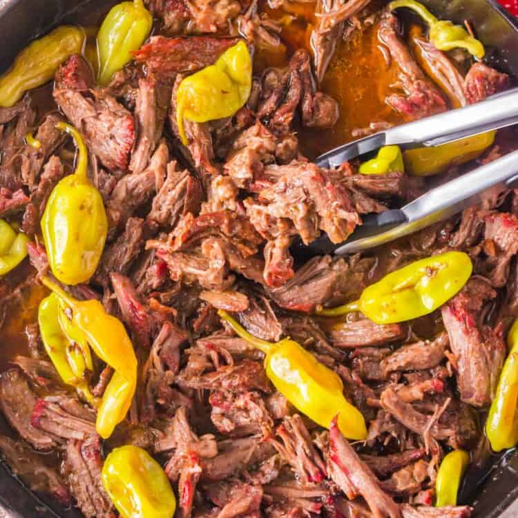 shredded Mississippi pot roast in a skillet with pepperoncini and gravy. Tongs are serving.