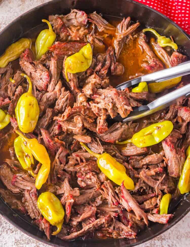 shredded Mississippi pot roast in a skillet with pepperoncini and gravy. Tongs are serving.