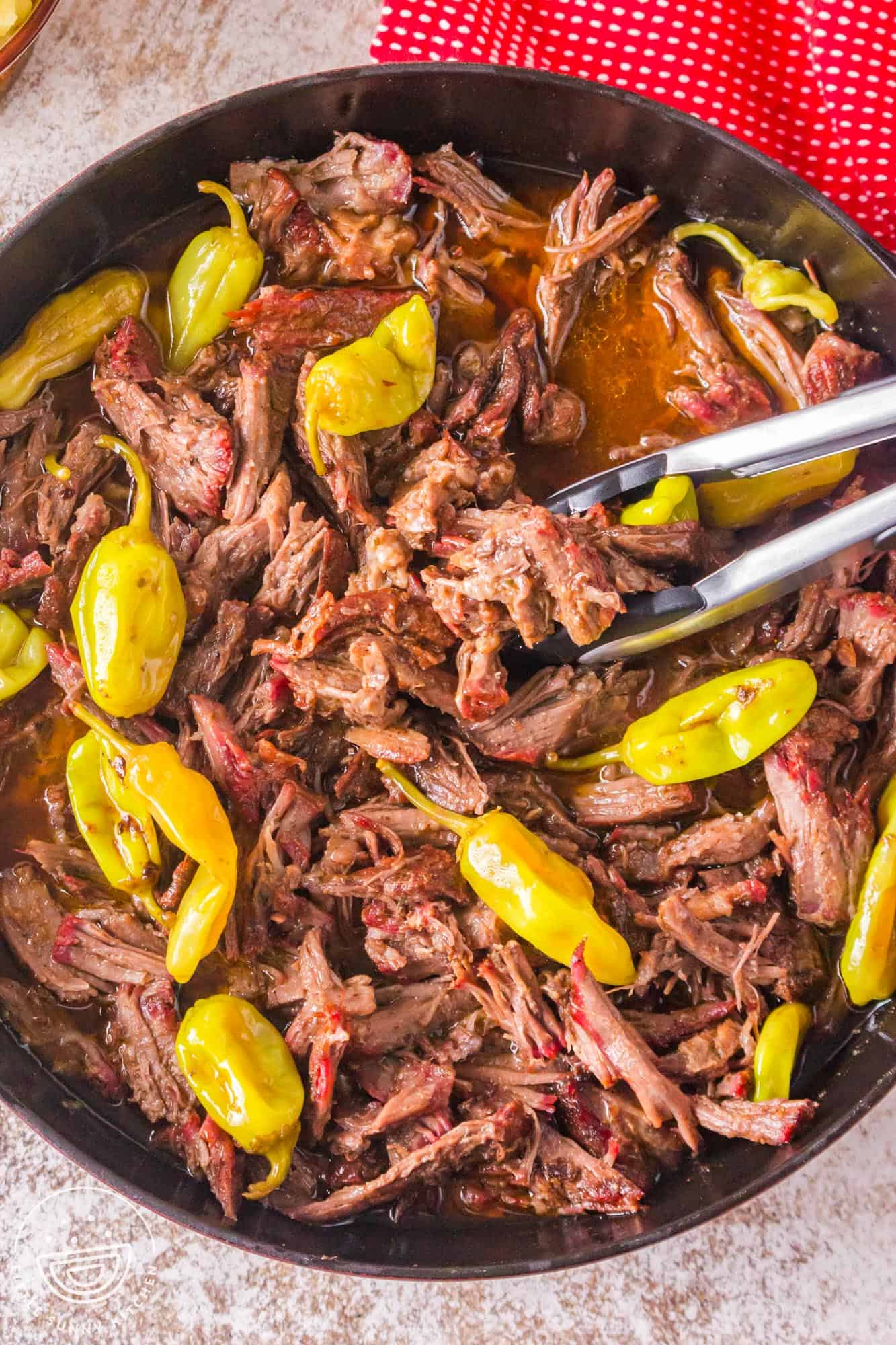 shredded Mississippi pot roast in a skillet with pepperoncini and gravy. Tongs are serving.