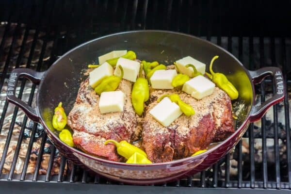 smoked chuck roast in a dutch oven inside of a smoker. The meat is topped with gravy mix, pats of butter, and pepperoncini.