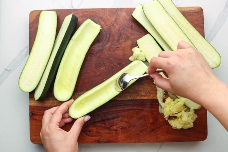 hands scooping seeds from halved small zucchini using a spoon on a wooden cutting board.