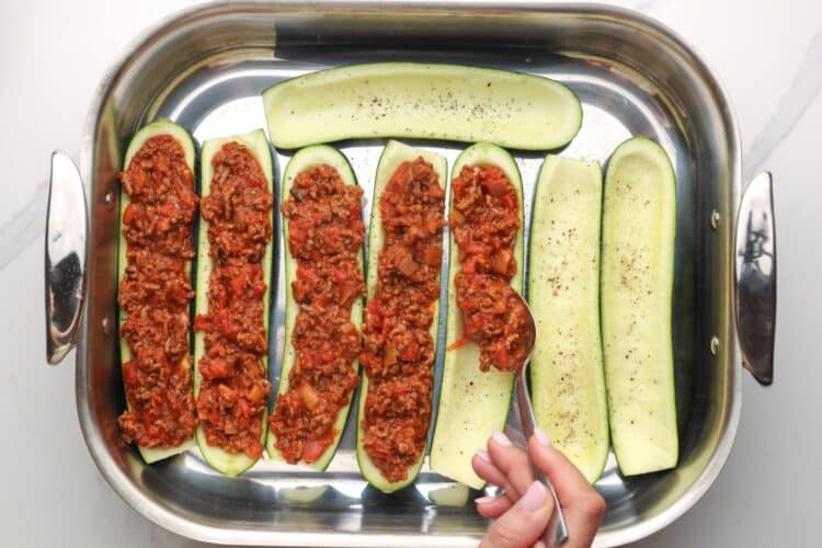 zucchini boats in a metal roasting pan, a hand is using a spoon to fill them with meat sauce.