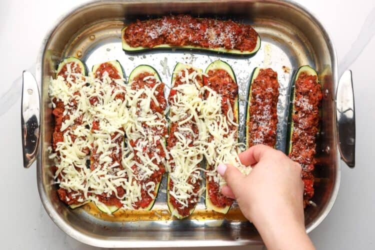 adding cheese to the top of ground beef zucchini boats in a roasting pan.