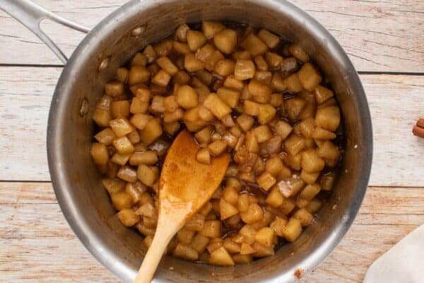 Cooked diced apples in a cinnamon sugar sauce in a metal saucepan, stirred with a wooden spoon.