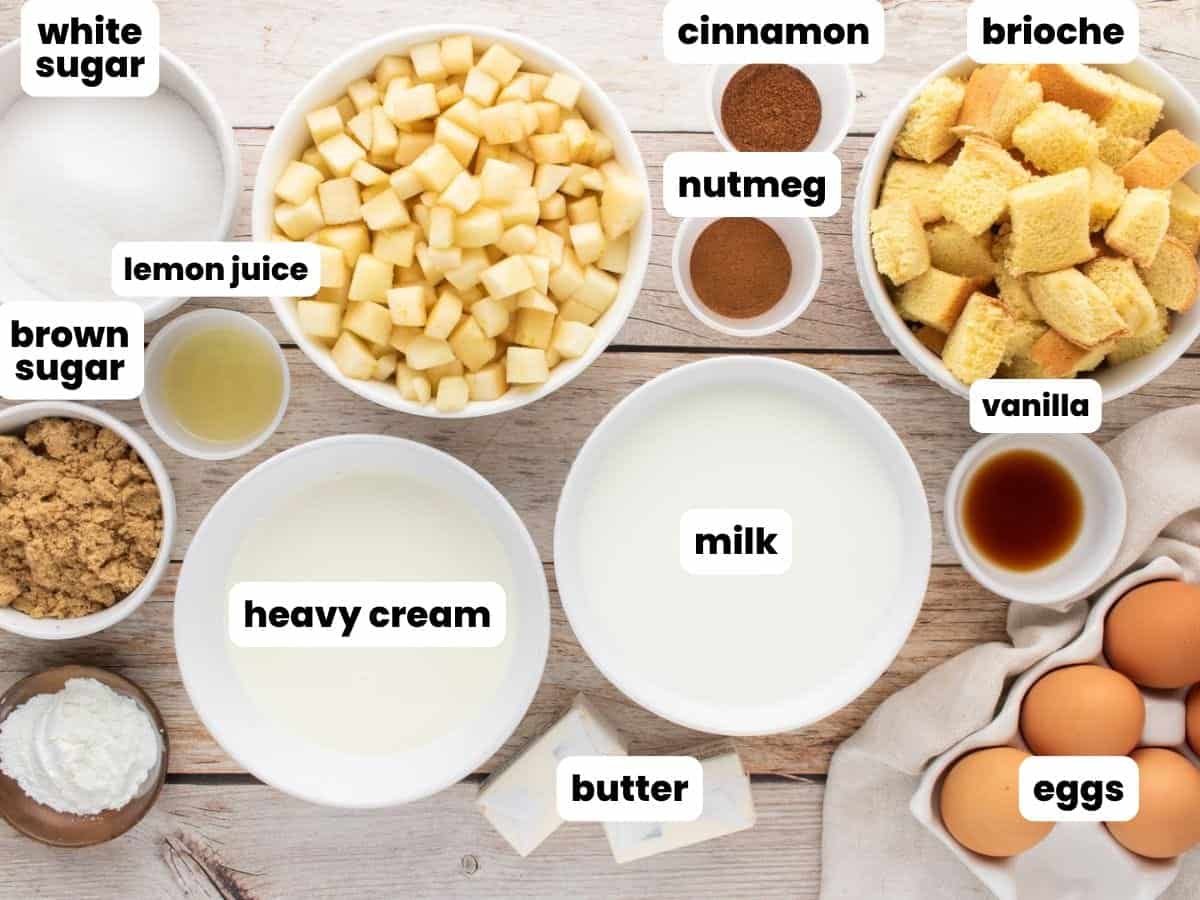Ingredients for apple bread pudding, measured into white bowls and arranged on a wooden table. Included are sugar, cinnamon, nutmeg, diced apples, brioche cubes, brown sugar, lemon juice, vanilla, heavy cream, milk, butter, corn starch eggs.
