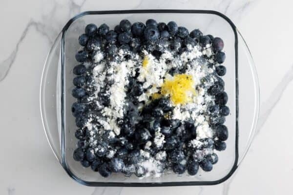 Fresh blueberries in a glass square baking dish, sprinkled with corn starch, sugar, and lemon zest.