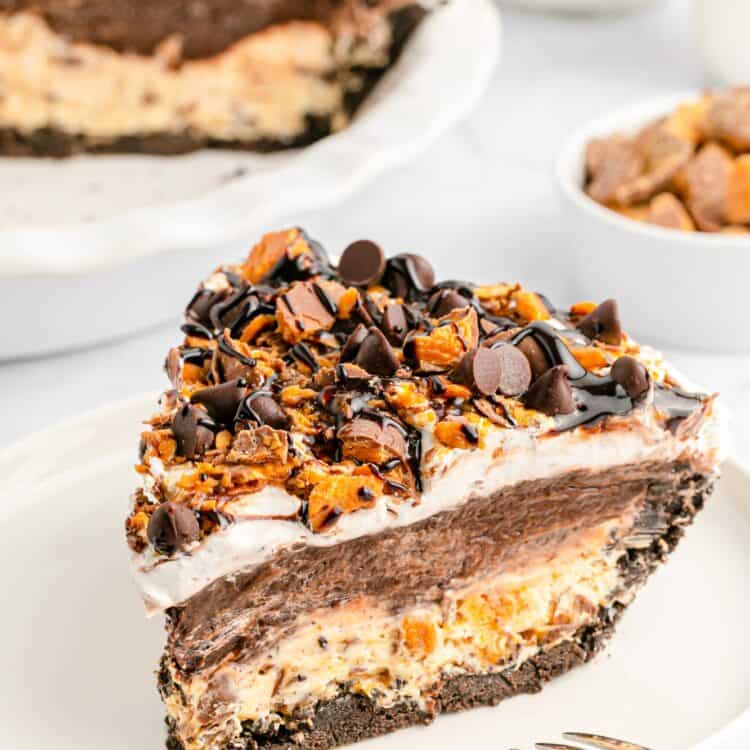 A close-up slice of no-bake chocolate Butterfinger pie on a white plate, showing layers of Oreo crust, peanut butter cream cheese filling, chocolate pudding, and whipped topping, all topped with chopped Butterfinger candy, semisweet chocolate chips, and chocolate syrup. The full pie is visible in the background, along with a bowl of chopped Butterfingers and glasses of milk.