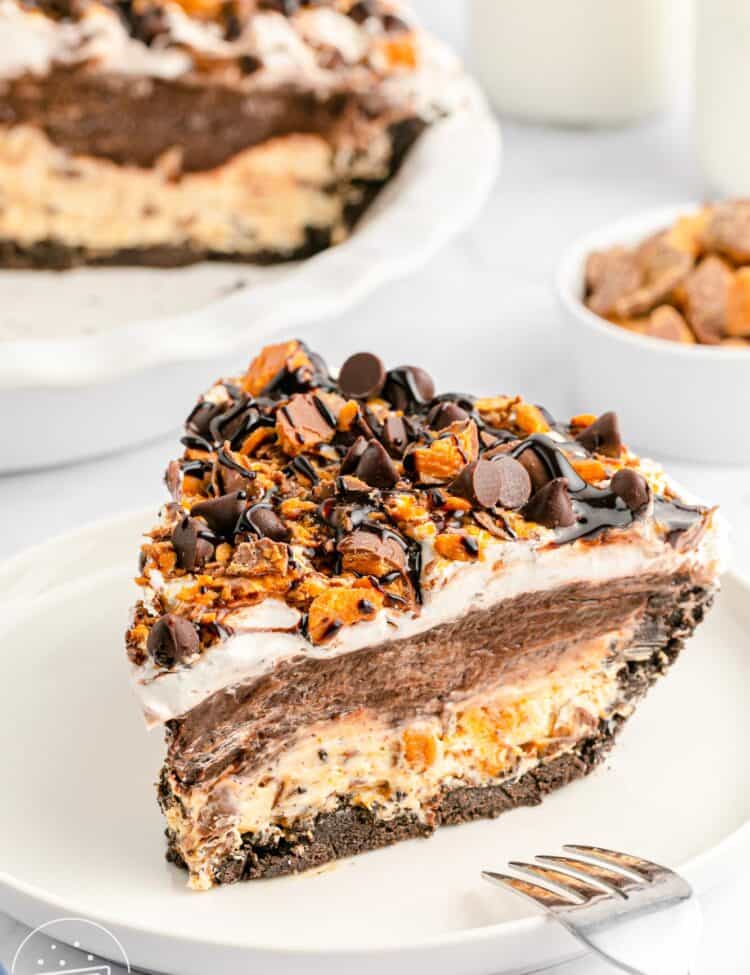 A close-up slice of no-bake chocolate Butterfinger pie on a white plate, showing layers of Oreo crust, peanut butter cream cheese filling, chocolate pudding, and whipped topping, all topped with chopped Butterfinger candy, semisweet chocolate chips, and chocolate syrup. The full pie is visible in the background, along with a bowl of chopped Butterfingers and glasses of milk.