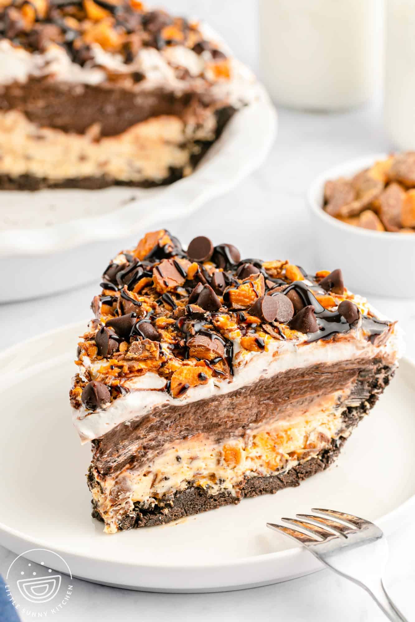 A close-up slice of no-bake chocolate Butterfinger pie on a white plate, showing layers of Oreo crust, peanut butter cream cheese filling, chocolate pudding, and whipped topping, all topped with chopped Butterfinger candy, semisweet chocolate chips, and chocolate syrup. The full pie is visible in the background, along with a bowl of chopped Butterfingers and glasses of milk.
