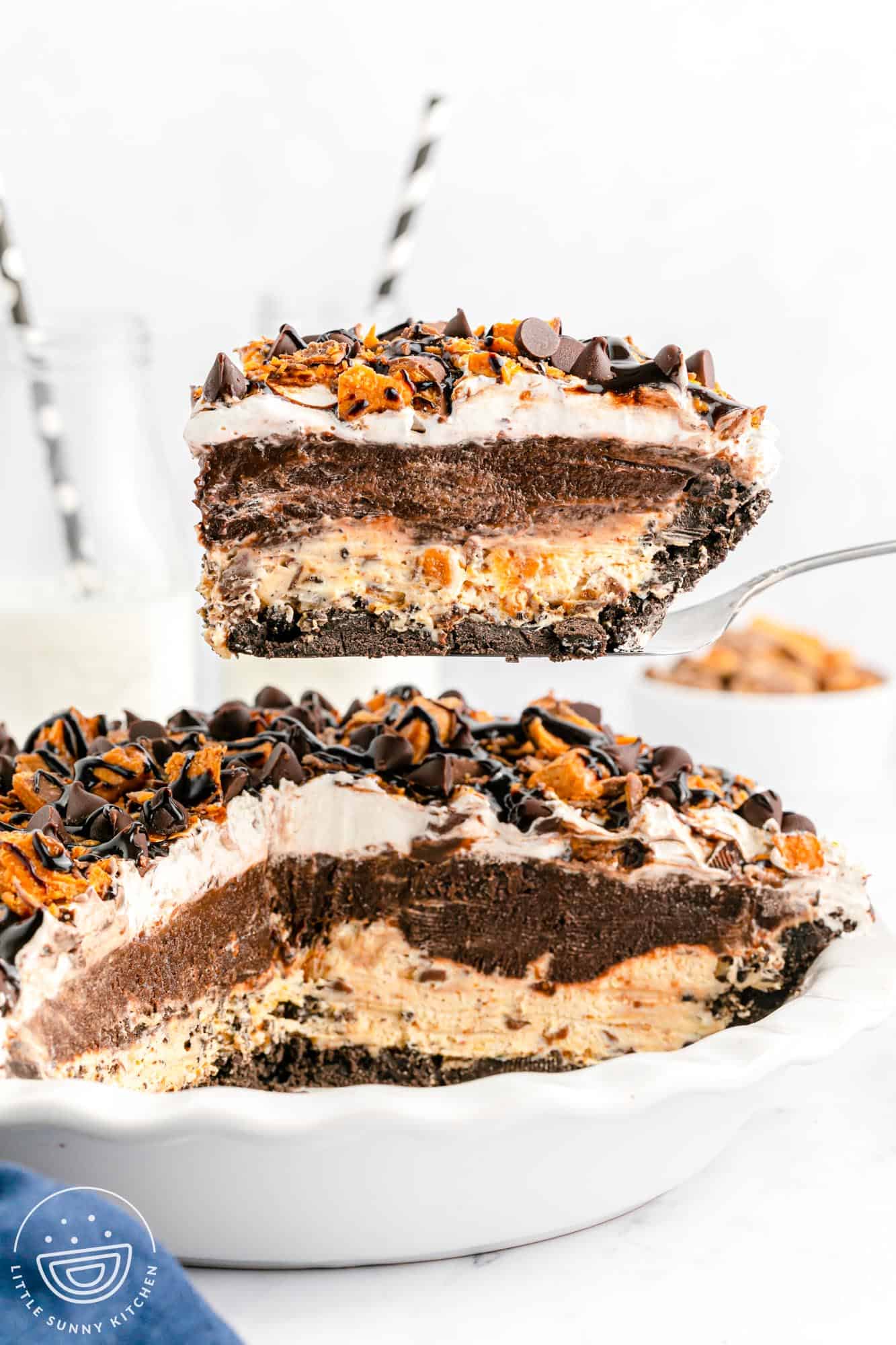 A slice of no bake chocolate Butterfinger pie being lifted from a pie dish, showing clear layers of Oreo crust, peanut butter cream cheese filling with chopped Butterfingers, chocolate fudge pudding, whipped topping, and a topping of more chopped Butterfingers and chocolate chips.